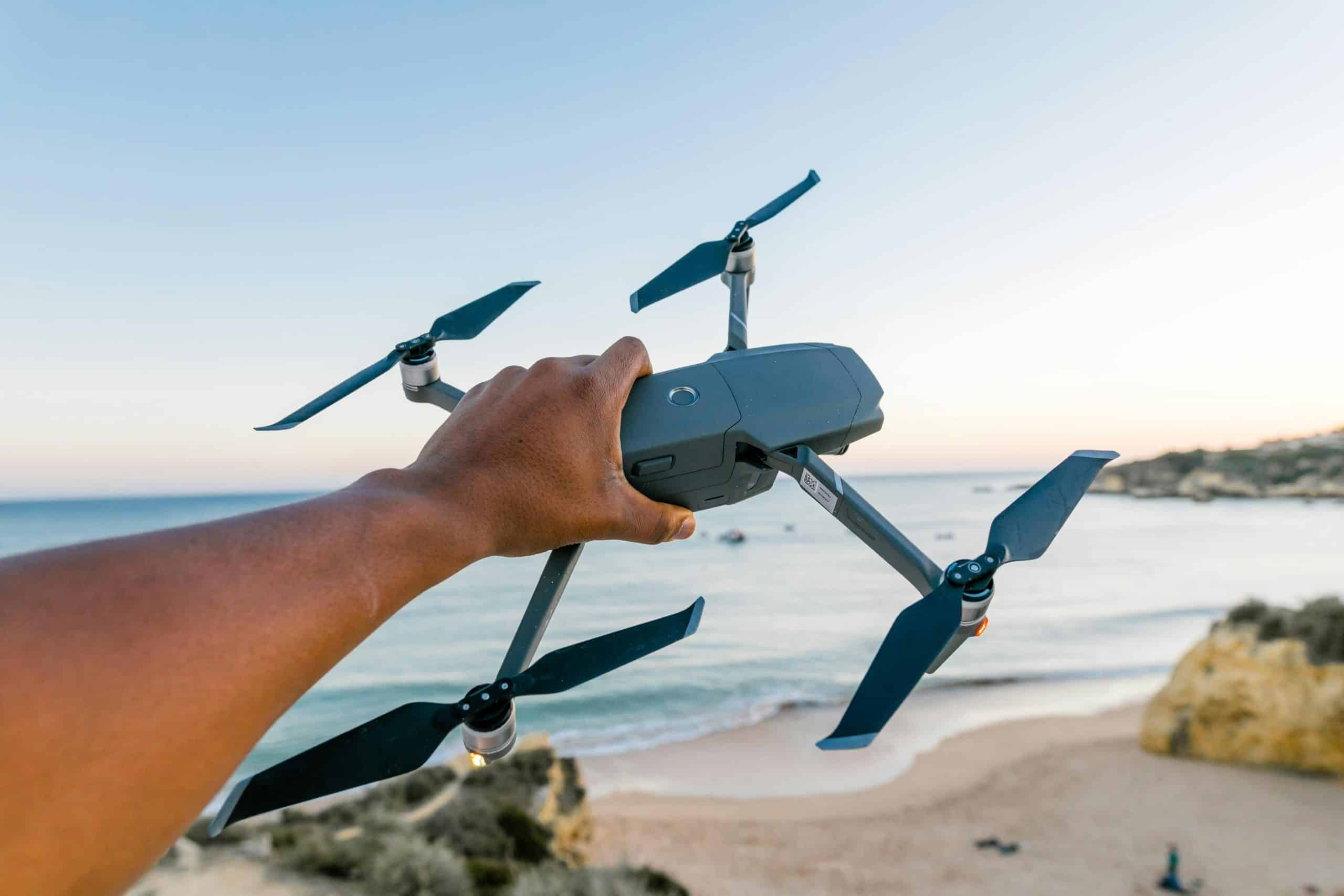 A hand holding a drone over a beach in Albufeira, Portugal, during sunset, showcasing technology and travel.